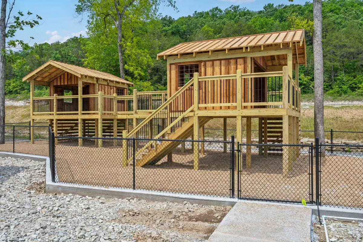 The image features wooden play structures within a fenced area, surrounded by lush greenery. Children are seen actively playing and exploring, while adults gather nearby for a family-friendly atmosphere. The vibrant outdoor environment emphasizes opportunities for imaginative play and family connection within nature.