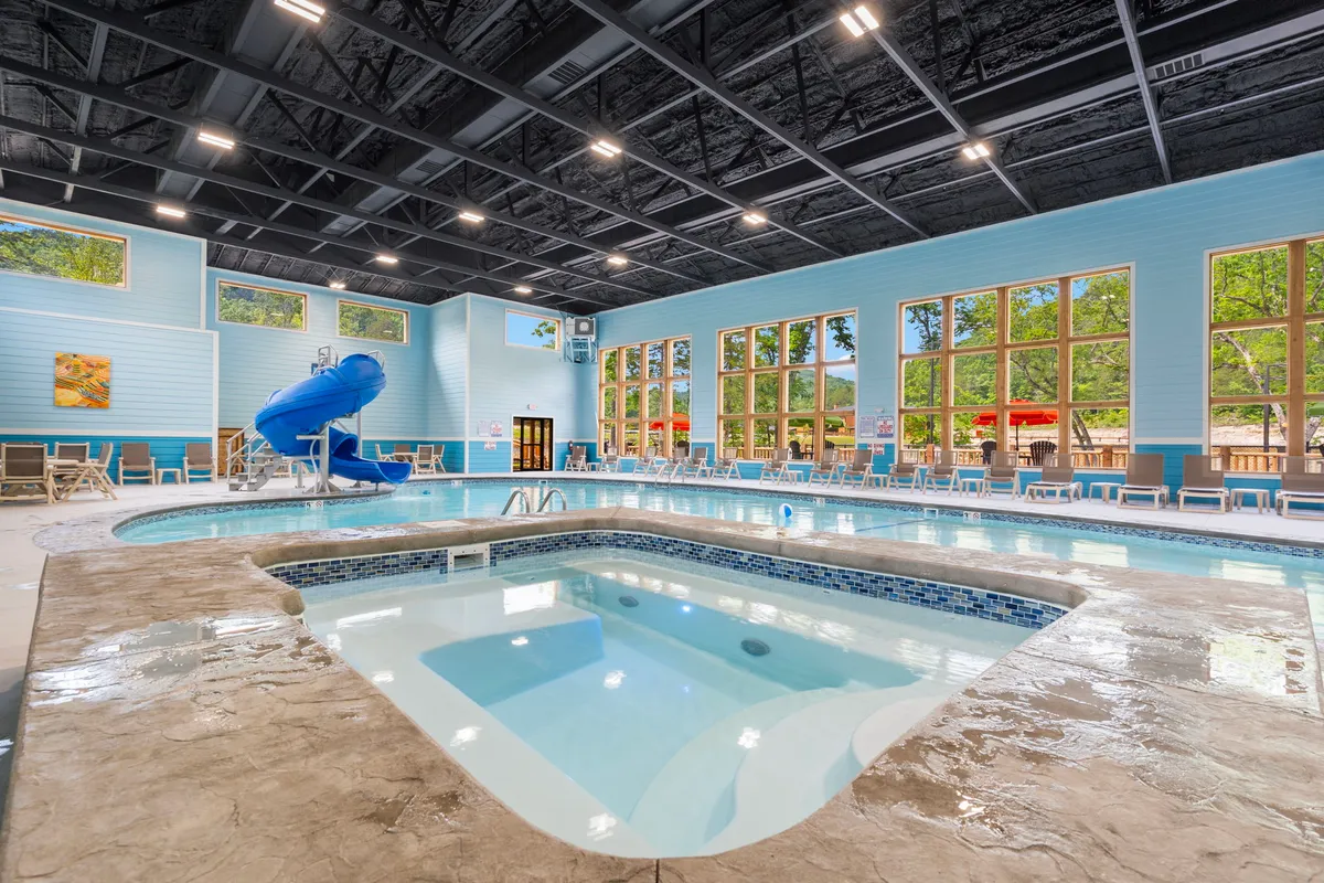 A bright, spacious indoor swimming pool area with a water slide, jacuzzi, and large windows letting in natural light. Children are seen playing on the slide and in the pool, while others relax in the jacuzzi and on lounge chairs nearby. The setting is energetic and family-friendly, with amenities designed for year-round fun.
