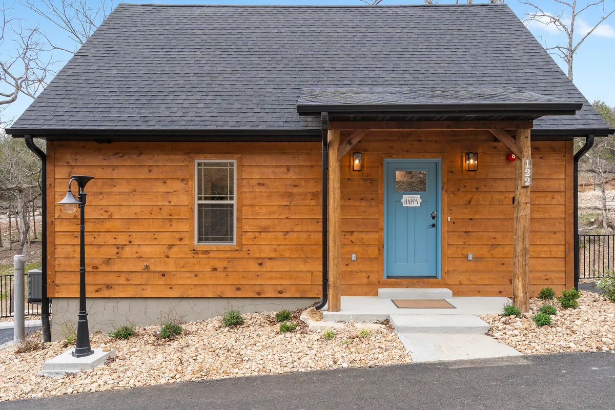 The image shows the inviting exterior of a charming wooden cabin in Hollister, Missouri. Its warm wooden façade and cheerful blue front door are accented by glowing lanterns and lush landscaping. A spacious front porch invites guests to relax and enjoy the tranquil wooded surroundings, perfect for gatherings with family or friends.