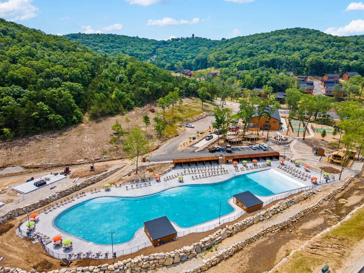 This photo displays a spacious outdoor swimming pool bordered by lounge chairs, a wooden deck, and cabana-style structures. Children and families can be seen enjoying the water, playing, and relaxing under the sun. The scene is set against a backdrop of greenery, exuding a serene yet sociable atmosphere.