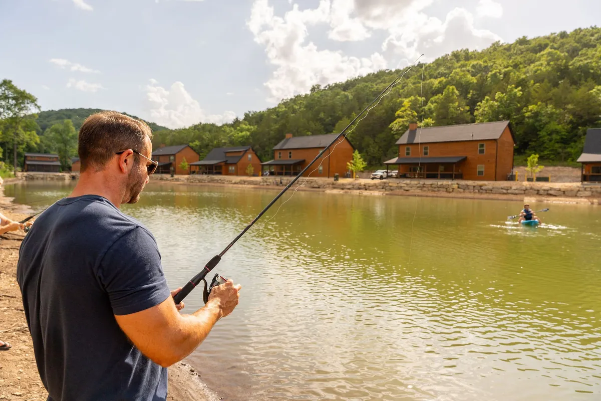 A tranquil lakeside scene featuring a man fishing beside reflective water, a kayaker paddling nearby, and wooden cabins along the shore. The image conveys peaceful recreation and the opportunity to connect with nature.