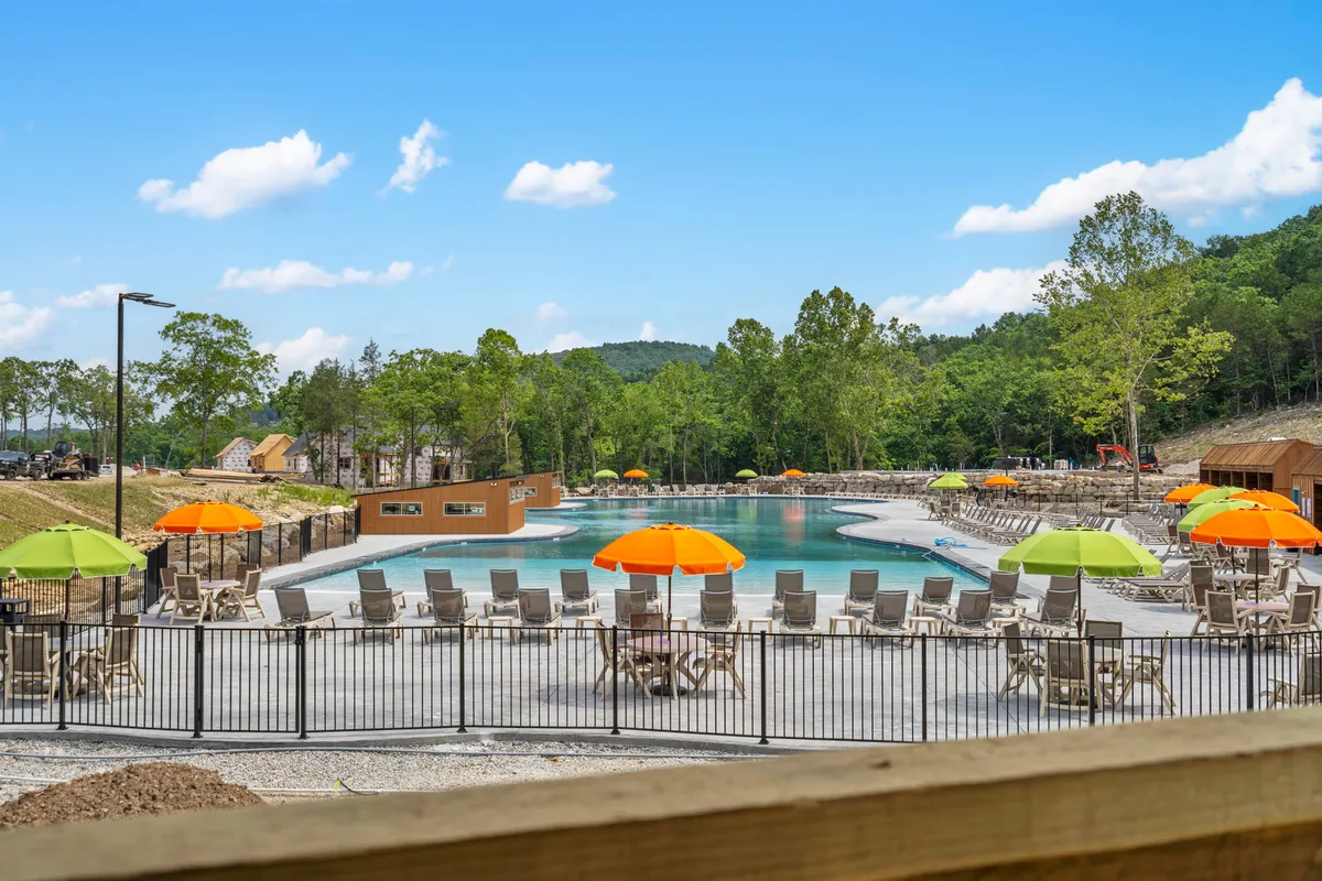 A sunny outdoor pool surrounded by colorful umbrellas, lounge chairs, and lush greenery. Families and children are seen swimming, sunbathing, and socializing, creating a cheerful, relaxed vibe. The scene evokes the feel of memorable summer days spent outdoors.
