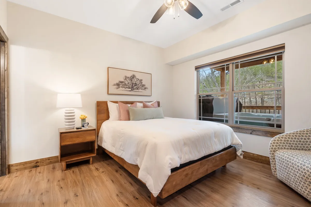 This interior image presents a comfortable bed with decorative artwork above, a nightstand with a lamp, and a window letting in soft, natural light. The space exudes tranquility and invites restful sleep or leisurely mornings with coffee.