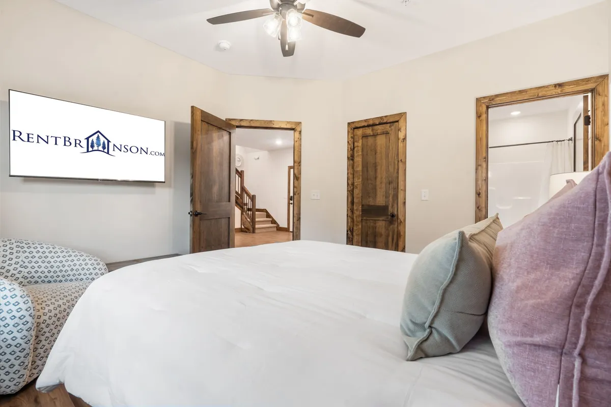 A cozy bedroom scene with a neatly made bed decorated with pillows, a TV across the room, and doorways leading to other spaces. There's also a staircase visible, hinting at the multi-level layout mentioned in the section.