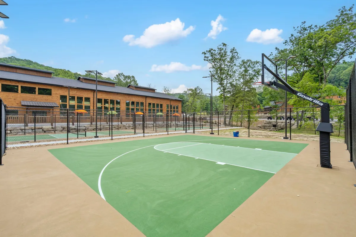 An exterior shot of a basketball court with a hoop, fenced area, and a scenic nature backdrop. The image also shows a building facade in the distance, creating a sense of community and outdoor recreation.