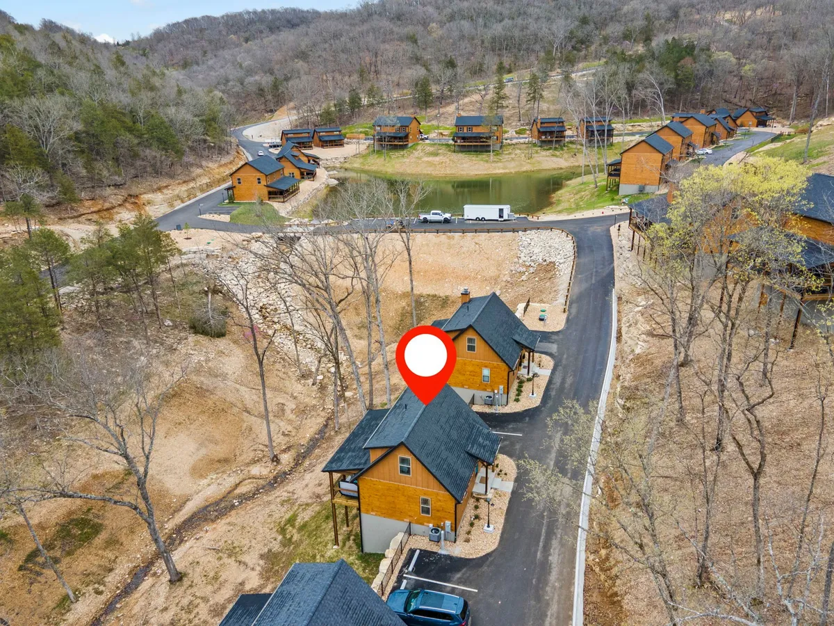 This image shows several wooden cabins nestled among tall trees, a circular pond reflecting the sunset, and a paved road winding through the property. The setting is tranquil, with ample green space and visible nature trails, conveying a sense of calm and immersion in nature.
