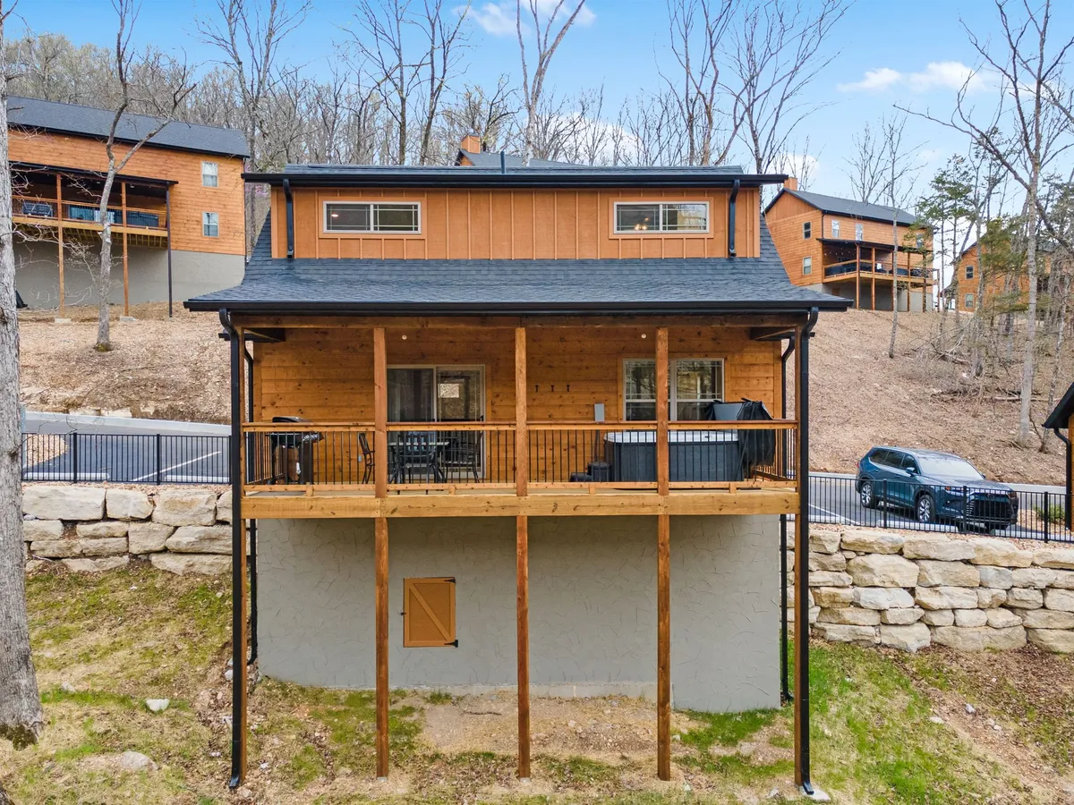 A wooden deck with comfortable seating overlooks a natural landscape, including trees and open sky. The deck is attached to a wood-sided cabin, and there is a visible hot tub, as well as a parking area nearby. The setting is cozy and relaxed, perfect for gatherings or quiet moments with nature.