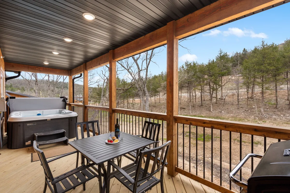 A tranquil exterior deck surrounded by trees features a private hot tub with steam rising from the water, an outdoor dining table, and cozy seating. The ambiance is peaceful and inviting, suggesting a place to relax and soak in nature, whether in the hot tub or with a meal outdoors.
