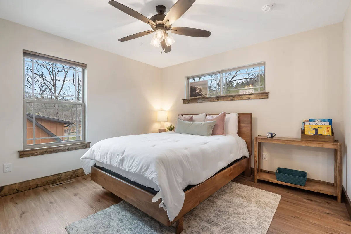A comfortable bedroom setup with a bed, nightstand, windows framing outdoor views, a ceiling fan, and small decorative touches, creating an inviting atmosphere for rest or quiet moments.